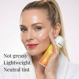A woman with blonde hair in a ponytail and a white robe applies a beige tinted sunscreen. Text on the image reads 'Not greasy Lightweight Neutral tint'.