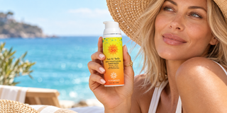 A woman in a straw hat and white swimsuit holds a bottle of Yunasence Sun Si Belle sunscreen near a sunny beach with turquoise water in the background.