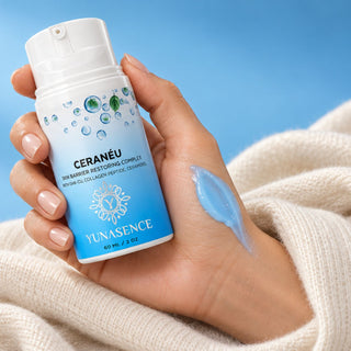 A hand holding a bottle of YUNASENCE CeraNeu RESTORING COMPLEX, with a blue swatch on the back of the hand and a soft cream-colored knit blanket against a bright blue background.