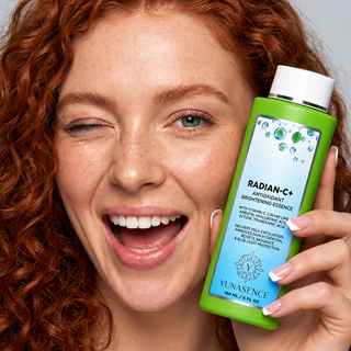 A close-up lifestyle shot of a smiling woman with curly red hair holding a bottle of YUNASENCE Radian-C+ Antioxidant Brightening Essence. The bottle is bright green with a white cap and a light blue label.