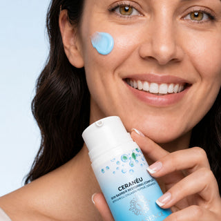 A close-up of a smiling woman holding a bottle of Ceraneu skin moisturizing cream while applying a dollop of the light blue cream to her cheek. The background is a clean, solid light blue.