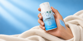 A hand holding a bottle of YUNASENCE CeraNeu RESTORING COMPLEX, with a blue swatch on the back of the hand and a soft cream-colored knit blanket against a bright blue background.