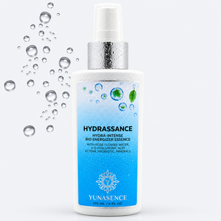 A white spray bottle of YUNASENCE Hydrassance essence with a silver pump and nozzle, decorated with blue water droplets and green leaves on the label. The background is a clean white with floating water droplets on the left side.