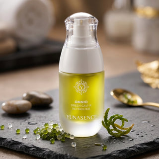 A bottle of YUNASENCE Omnio Green Cavi-R Retinol Krème centered on a dark slate tray with green beads, a gold spoon, and blurred natural props in the background.