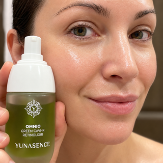 A close-up of a woman holding a bottle of Yunasence Omnio Green Caviar Retinolix skincare serum next to her cheek, showing the product in use.