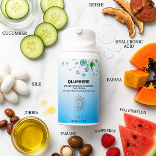 A bottle of Yunasence Qlumiere Bio Restorative Cucumber Day Cream is centered on a white marble background, surrounded by ingredient slices and droplets labeled cucumber, reishi, hyaluronic acid, silk, papaya, watermelon, raspberry, jojoba, tamanu, and macadamia. The bottle has a white cap and a blue-to-white label with green and blue droplet graphics.