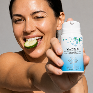 A smiling woman holds a white and blue bottle of YUNADENCE QLUMIERE Bio Restorative Cucumbers Day Cream, with a cucumber slice held between her teeth against a neutral gray background.