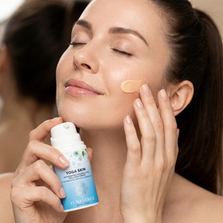 A woman with closed eyes applies a dollop of Yoga Skin Face cream to her cheek while holding the bottle in her hand.